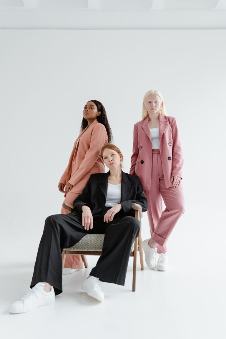 Three women in suits pose. Two are standing, one in an orange suit and the other in a pink suit, behind a woman sitting. The woman who is sitting is wearing a black suit