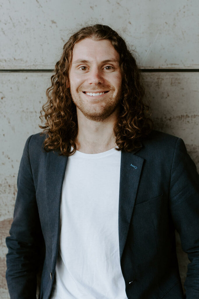 Image shows Jono Naef in a black blazer and white tshirt smiling toward the camera